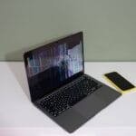 Uszkodzony ekran laptopa z kolorowymi liniami obok żółtego smartfona